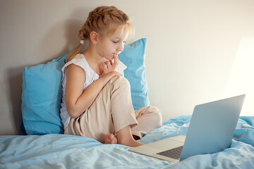 A cute girl sitting on the bed is watching something on a laptop. The concept of education.