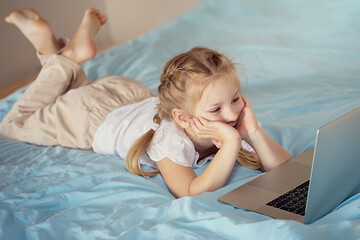 A cute girl sitting on the bed is watching something on a laptop. The concept of education.