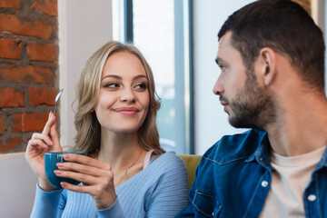cheerful blonde woman holding coffee cup and teaspoon near blurred boyfriend
