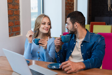 happy couple with coffee cups looking at each other near blurred laptop in restaurant