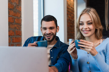 cheerful couple with coffee cups looking at blurred laptop while spending time in cafe