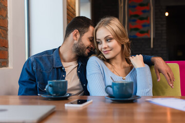 happy man embracing girlfriend near blurred coffee cups, smartphone and laptop in restaurant