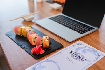 delicious sushi rolls, menu and blurred laptop with blank screen on table in sushi bar