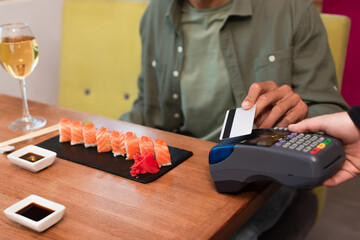 cropped view of blurred man paying through credit card reader near sushi rolls and wine glass