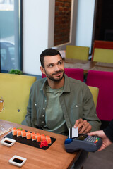 smiling man paying with credit card through payment terminal near tasty sushi