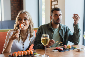 man with closed eyes enjoying delicious sushi near smiling girlfriend