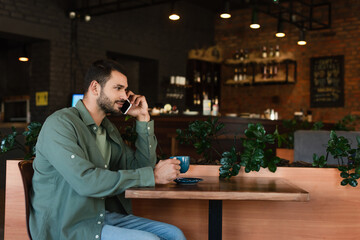 side view of young man talking on smartphone while sitting in cafe with coffee cup