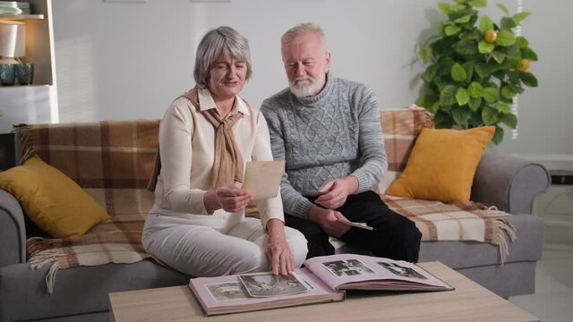 elderly life and family memories, gray-haired cute aged couple looking at old photos in album at home