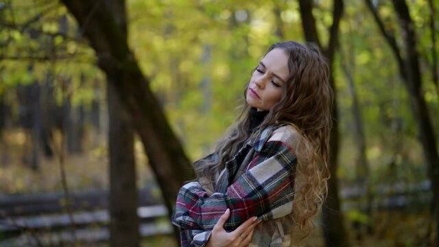 portrait of a woman with curly hair and a plaid coat in an autumn park. she hugged herself and closed eyes. the camera is moving