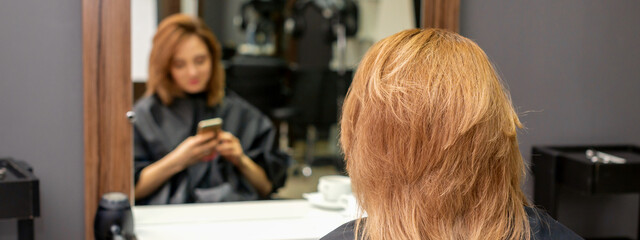 Beautiful young red hair woman using her smartphone and texting sitting in front of a mirror waiting to visit a hairdresser