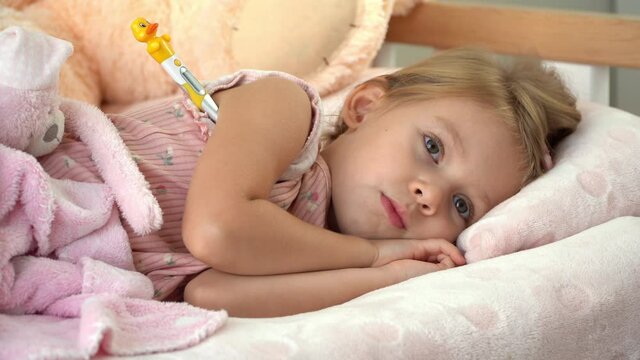 Sick little child in bed with thermometer looks at camera and coughs. Temperature measurement in illness. Treatment for flu and cold