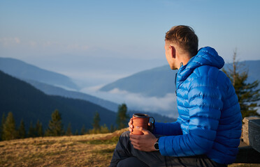 Hiking in the mountains. Guy sitting outdoors on mountain meadow at sunny morning, looking at very...