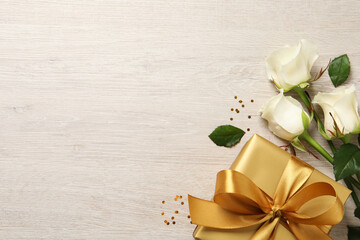 Golden gift box and beautiful roses on wooden background, flat lay. Space for text
