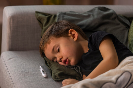 Adorable Little Child Boy Sleeping In The Bed Facing The Camera. Young Boy Is Sick At Home And Sleeping In His Bed With A Thermometer. Sick Boy Sleep Under Blanket With Thermometer