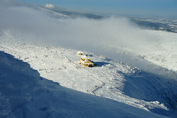 Aerial view of mountain cottage above clouds covered with snow, beautiful Giant Mountains on winter sunny day, Poland