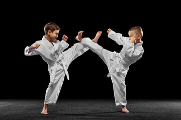 Two little kids, boys, taekwondo athletes training together isolated over dark background. Concept...