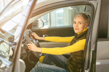 People, driving, transport concept. Portrait of middle age caucasian blond smiling woman sitting in the electric car at driver seat and looking at the camera.