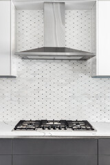 A stainless steel stove top and range hood surrounded by white and grey cabinets, tile backsplash, and granite countertop.