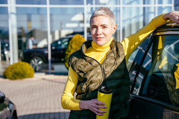 Positive woman in casual wear standing near white auto outdoor. Female customer rent a new car...