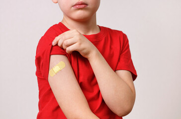 Little boy in red t-shirt shows his shoulder With A Patch After Being after vaccination. Children hand with Adhesive plaster after Injected With The Covid-19 Vaccine.