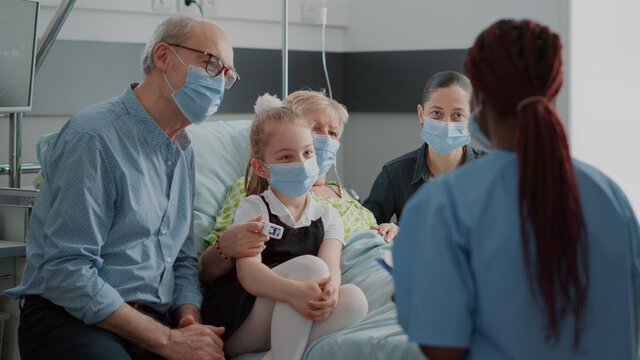 Nurse Giving Recovery Advice To Patient And Visitors With Face Mask In Hospital Ward. Medical Assistant Helping With Medication At Checkup Visit, Talking To Sick Woman And Family During Pandemic.