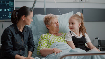 Ill pensioner enjoying visit from niece and daughter in hospital ward bed. Woman and little girl...