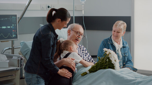Family Visiting Elder Patient With Illness In Hospital Ward Bed. Mother And Child Bringing Flowers To Grandpa At Visit In Intensive Care Room. Visitors Giving Comfort To Old Man. Handheld Shot