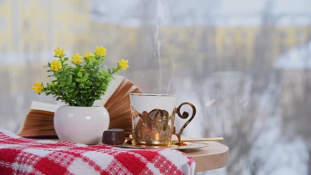 Golden coffee cup with smoke, book and vase with flowers on table in cafe. Winter time, It snows outside. Coziness atmosphere