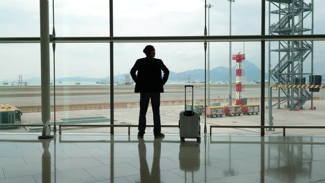 Man make step to window and stand against, look out to apron and empty taxiways. Passenger take breath after long international flight, rest at arrival terminal passage