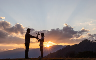 Silhouette Christian father holding Christian cross teaching his daughter to pray outside on...