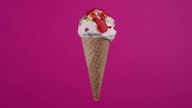 Yummy vanilla ice cream in waffle cone and pouring strawberry topping. Sweet dessert decorated with colorful sprinkles, rotating soft cream, gelato icecream scoop over pink background