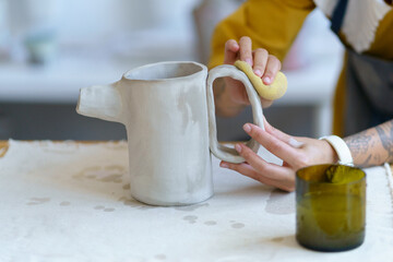 Creative young woman shaping clay jug in studio. Female artist with tattoos on hands work on...