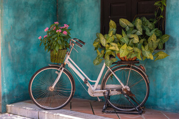 Fototapeta na wymiar White vintage bike with basket full of flowers next to an old building in Danang, Vietnam, close up