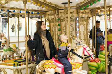 Mom and son are having fun on the merry-go-rounds. Child boy rides a carousel horse, amusement.	
