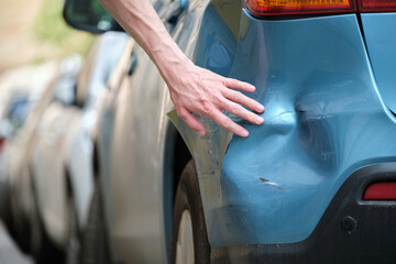 Driver hand examining dented car with damaged fender parked on city street side. Road safety and...