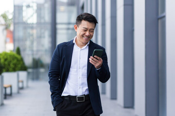 asian male freelancer walking near business center holding phone, smiling reading news, successful...