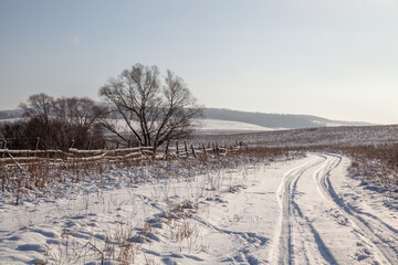 Fototapeta na wymiar Winter trail through snowy hills. Trees grow along the road behind a wooden fence.