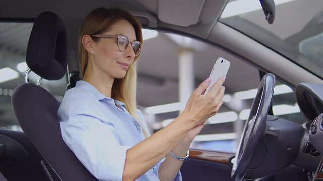 Beautiful self-confident blond woman in stylish shirt sitting on driver seat of her respectable car and scrolling web-pages on smartphone, doing shopping online, checking bank account