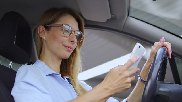 Smiling beautiful woman with blond hair in stylish eyeglasses sitting on driver seat holding hand on steering wheel, using navigation app on smartphone to find proper way, maps online, gps navigation