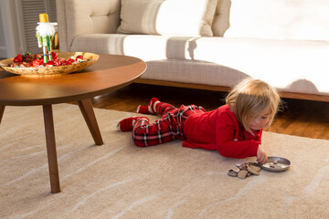 Cute blond toddler girl in red pyjamas lying down on carpet playing with small rocks in sunny living room