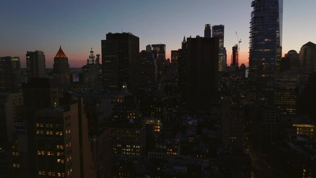 Fly above city at dusk. High rise buildings with lighted windows. Silhouettes against colourful sky. Manhattan, New York City, USA