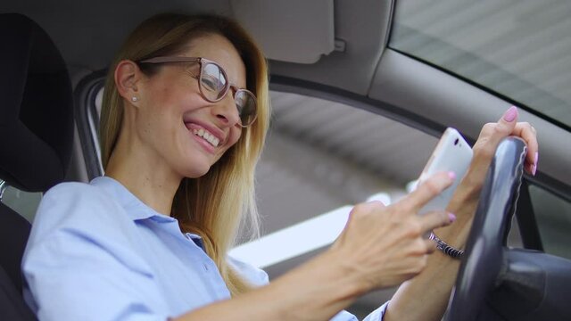 Extremely happy positive blond woman in stylish eyeglasses sincerely laughing reading good news or watching funny videos or photos on her smartphone, receiving message during driving