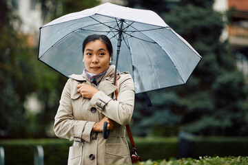 Asian businesswoman feels cold while walking with umbrella in park during rainy day.