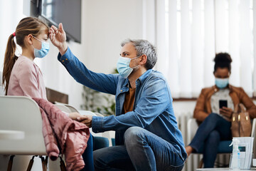 Obraz premium Worried father checks daughter's forehead for fever in waiting room at doctor's office during coronavirus pandemic.