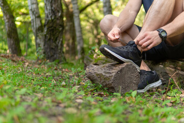 Man tying his shoelaces in the park outdoors, around the forest, oak trees green grass young enduring athletic athlete. run sport nature, lifestyle outdoor legs marathon, trees man. Summer body cross