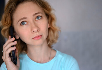 Close-up portrait of young caucasian woman talking on cell phone at home.The blue-eyed blonde looks up thoughtfully. She is wearing a blue T-shirt.