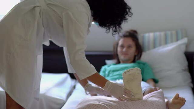 Smiling female doctor checking on a girl with a broken leg in plaster cast, female patient laying down in a hospital bed in pajamas