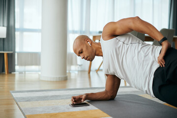 Black athletic man in side plank pose uses smart phone while exercising at home.