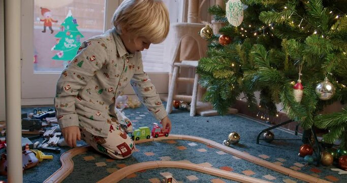 Cheerful cute child boy playing near Christmas tree with toy cars on railway wooden road at home on living room. Concept of carefree childhood on winter holidays