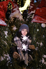 Outdoor young beautiful happy Face of smiling girl posing. Christmas tree Happy New Year handling sparklers lights among trees with white noses and christmas hats